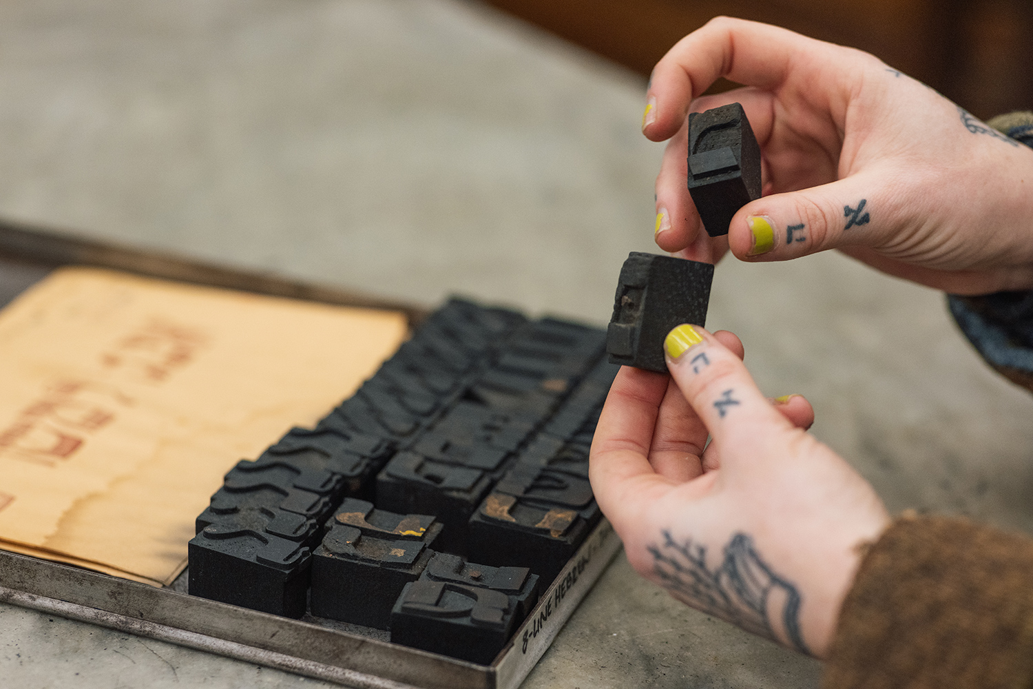 Two hands hold up two small blocks of Yiddish type. Below them is a small box full of blocks of Yiddish type.