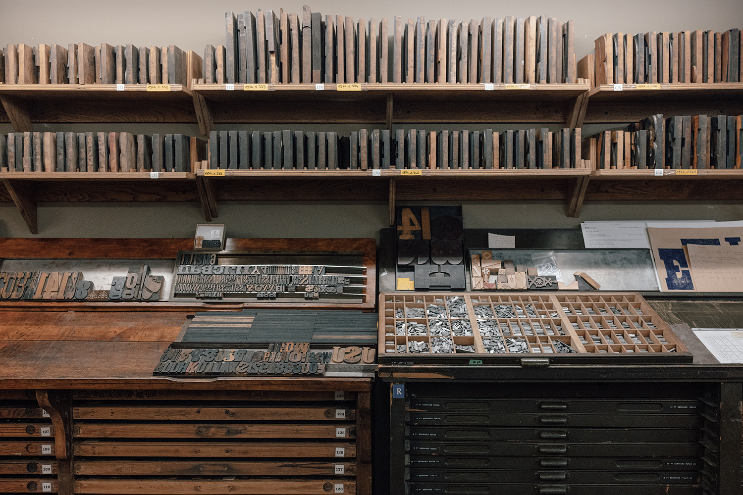 Two shelves against a wall hold large wooden blocks of type. In the foreground are two desks, each with several narrow drawers, which hold smaller type sizes. On top of the desk are also several trays of small tyoe.