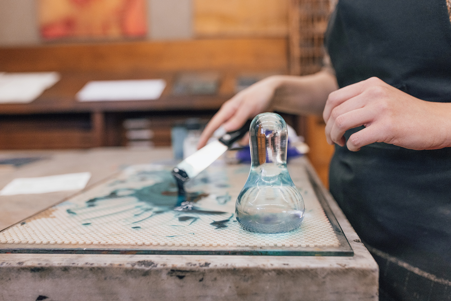 On a table with green ink is a glass pestle. A hand holds a knife with a wide blade that scrapes the ink along the table.