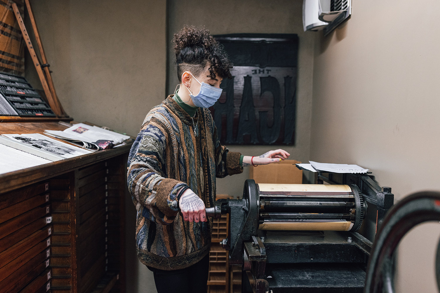 A woman operates a galley proof press. Next to her is a desk with several narrows drawers containing type.