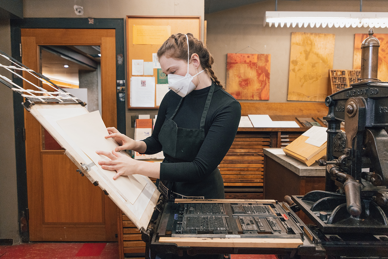 A woman sets a large piece of white paper in place on an upright tray. She is standing in front of an iron hand press with a metal tray containing inked type.