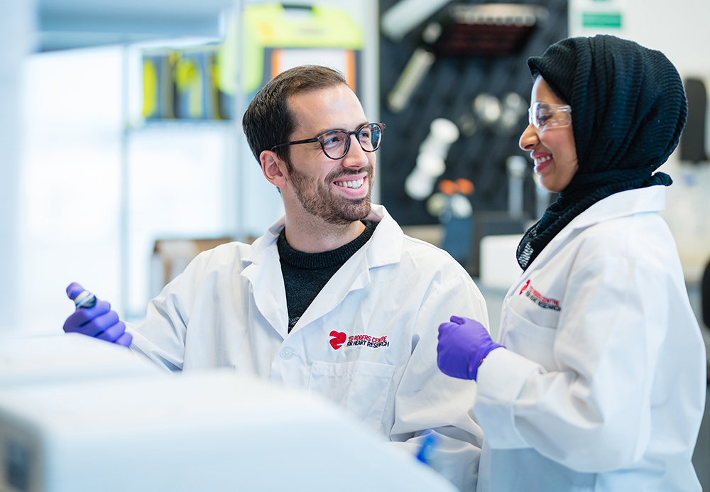 Two researchers wearing white lab coats, protective eyewear and blue disposable gloves, working and smiling. The female researcher on the right is wearing a hijab.