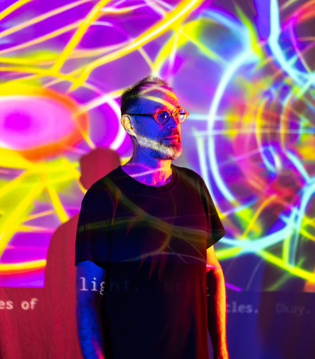 David Rokeby in glasses and a black T-shirt, standing in front of a screen, with multiple colours in various patterns projected on the screen