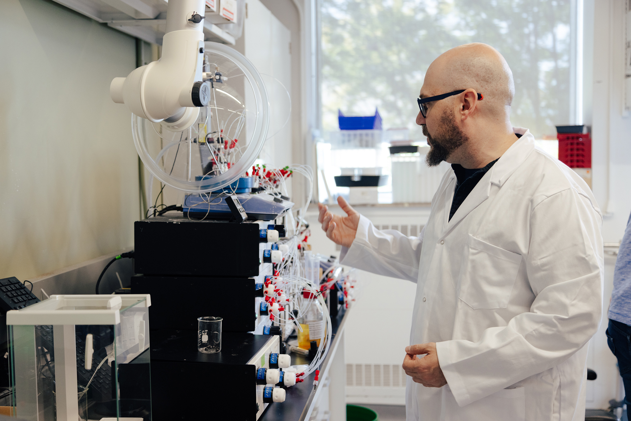 A bald man in a lab coat with black glasses and a beard looks at a machine with many wires emerging from it.