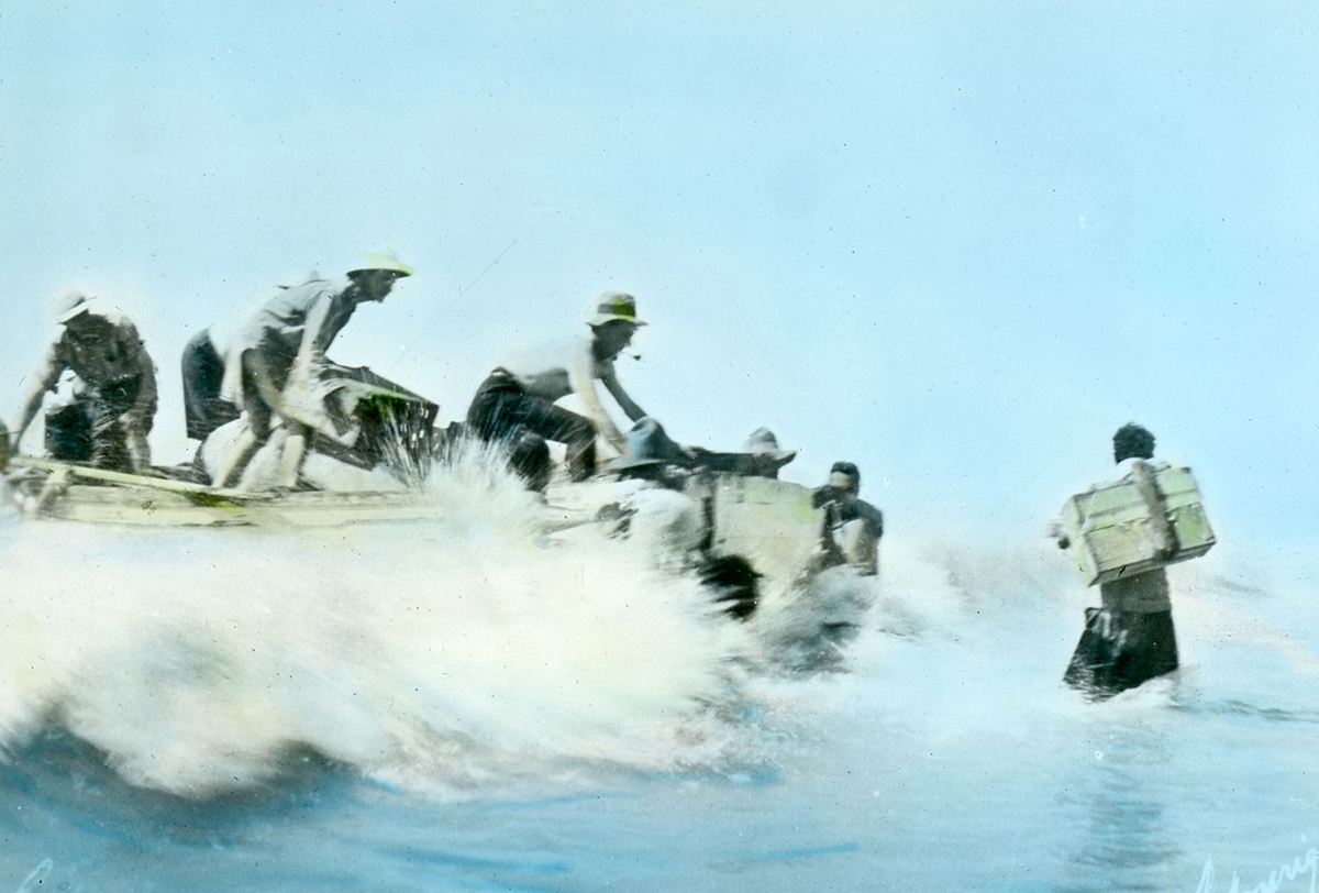 A heavy surf crashes against a whaleboat while scientist are moving crates of equipment off the boat. A couple of Nyangumarta people are helping the scientists carry the crates.