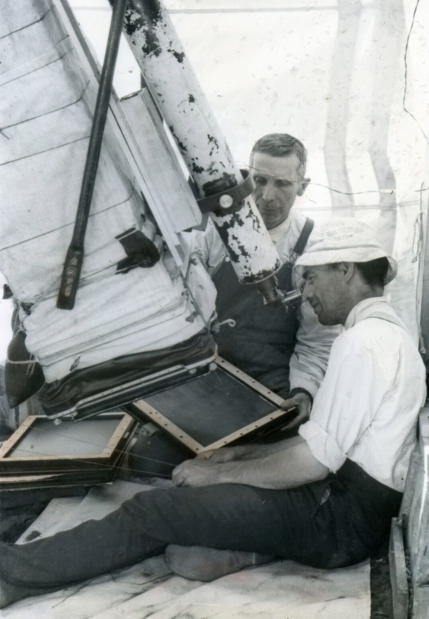 Black and white photo of astronomers Clarence Chant and Reynold Young kneeling and sitting, respectively, on the ground. Young is looking through the eyepiece of a long cylindrical-shaped camera positioned perpendicular to the ground and attached to a square tube covered with a cloth-like material. Chant is holding a black rectangular board underneath the tube.