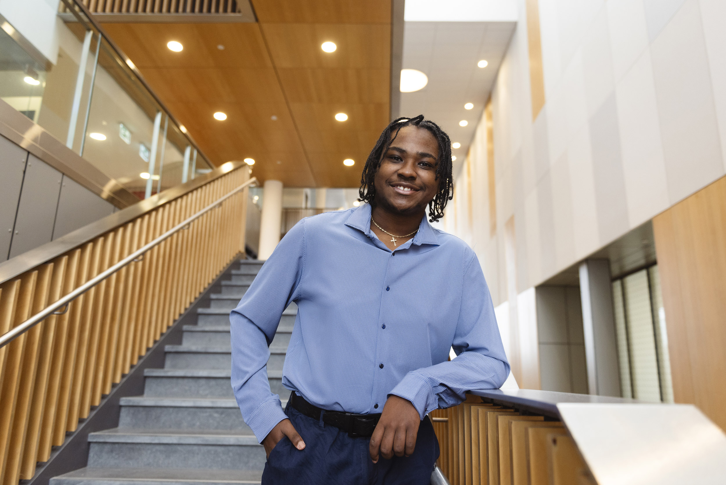 De-Mario Knowles, in a light grey-blue, long-sleeved button up shirt and navy blue trousers, standing on a straight staircase, with his elbow resting against the wide, metal surface running along the top of a wooden stair railing. He is wearing dreadlocks and a thin chain necklace with a cross.