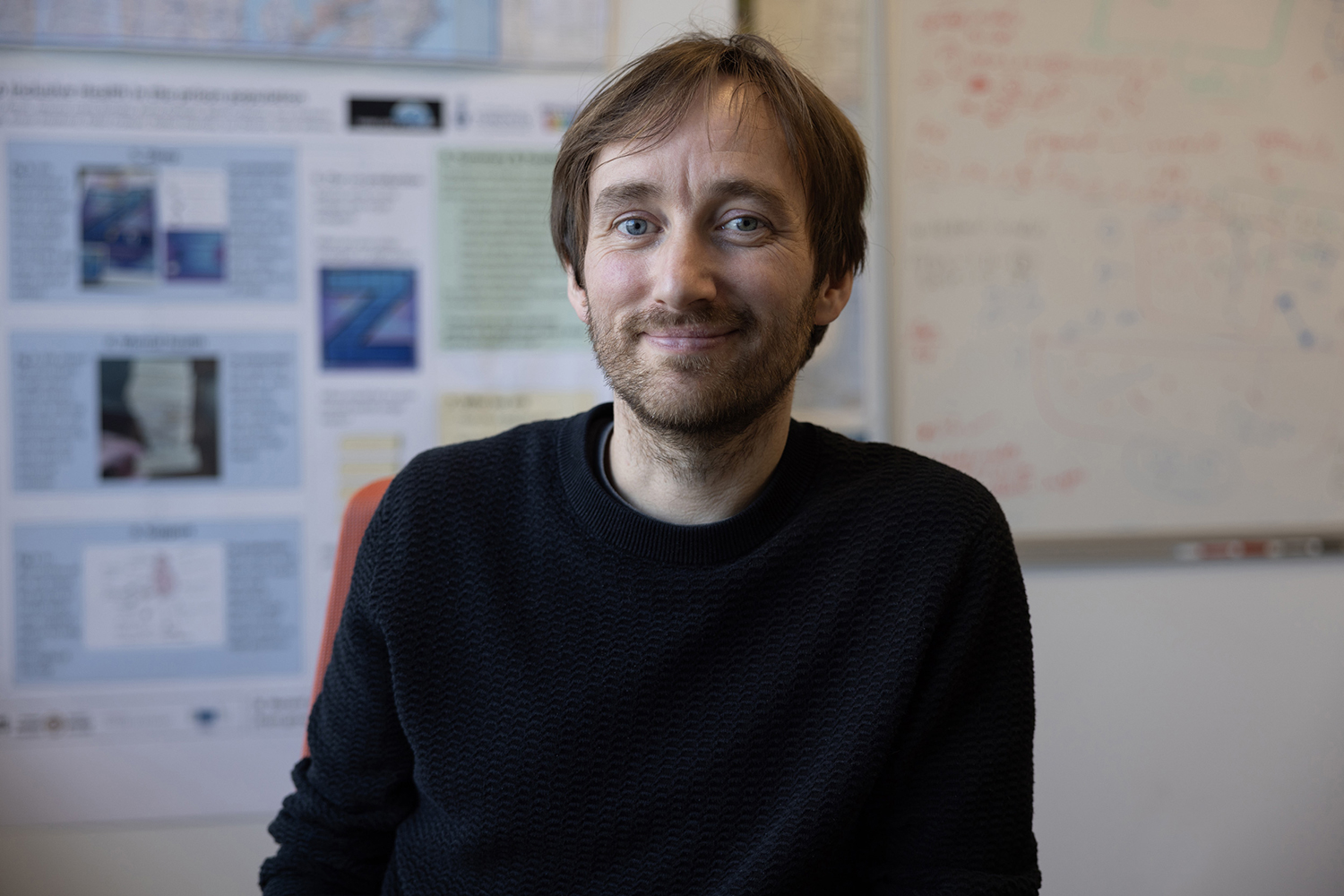 Phil Heron in a black sweater, sitting with a presentation poster and a white board with notes out of focus in the background behind him