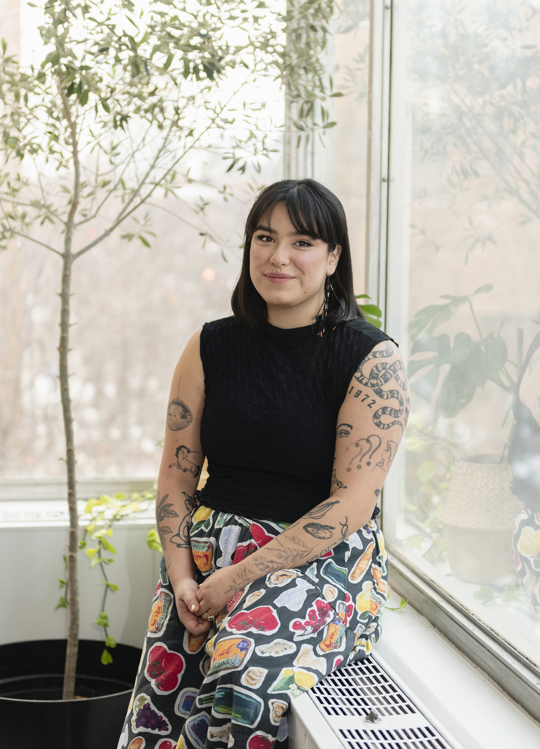 Riley Yesno is sitting on a radiator close to a corner, next to high windows. She is wearing pants with colourful illustrations of fruits and vegetables and a sleeveless black top, revealing tattoos that decorate her arms and shoulder.