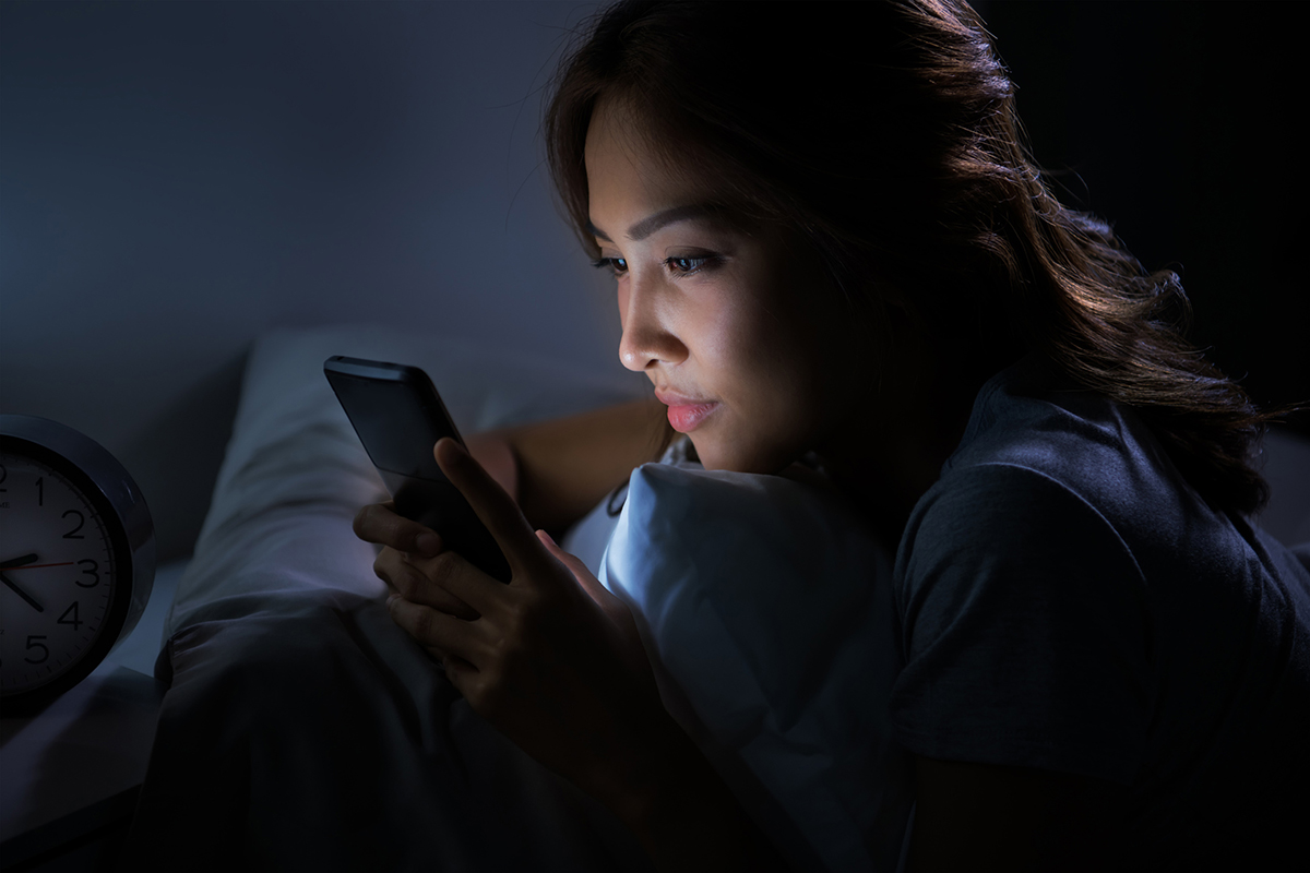 A young woman is looking at her cellphone in the dark, while lying on her front in bed