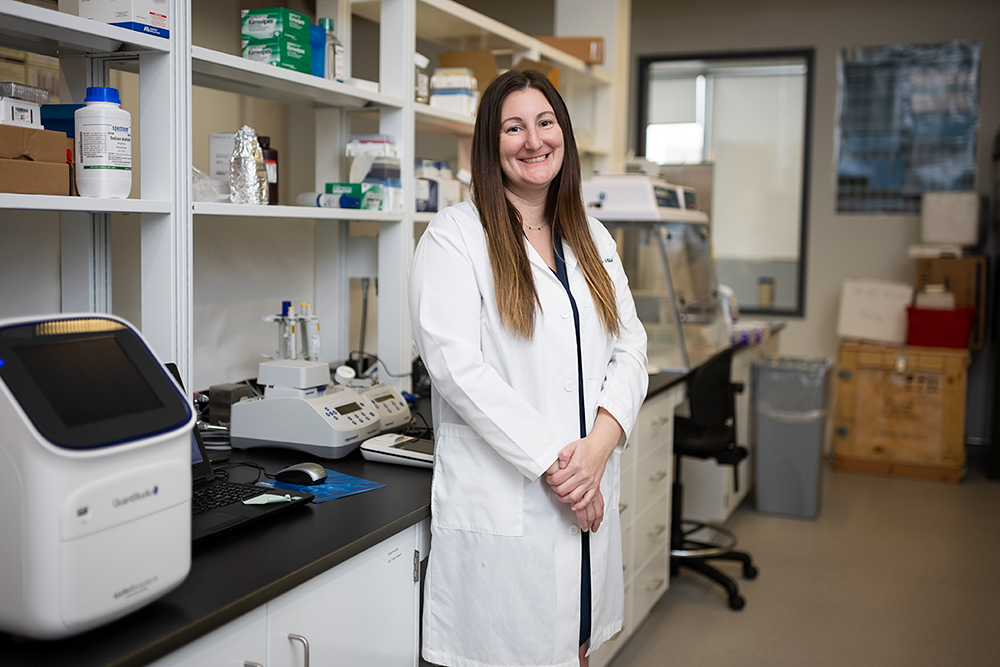 Nicole Novroski, a professor of forensic science, in a white lab coat, standing in front of shelves and desks in a science lab