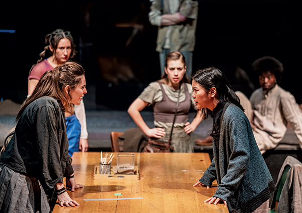 Students acting in a scene from The Trials. Two students appear to be in confrontation across a wooden table, with other students in the background, watching the pair.