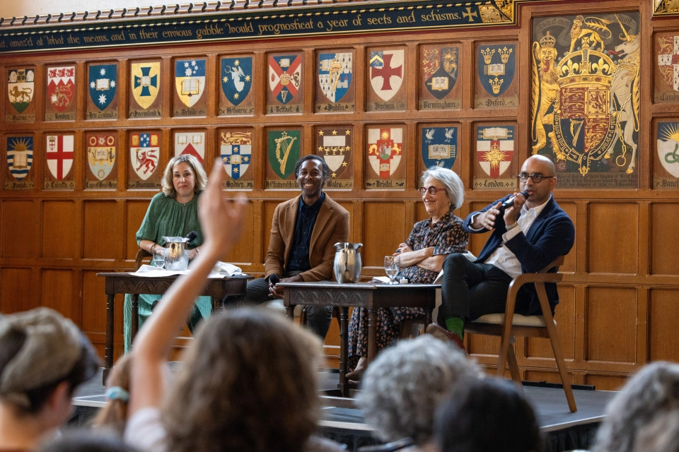 Four panellists are seated on a stage. Professor Randy Boyagoda is speaking into a microphone, and an audience member has one hand raised.