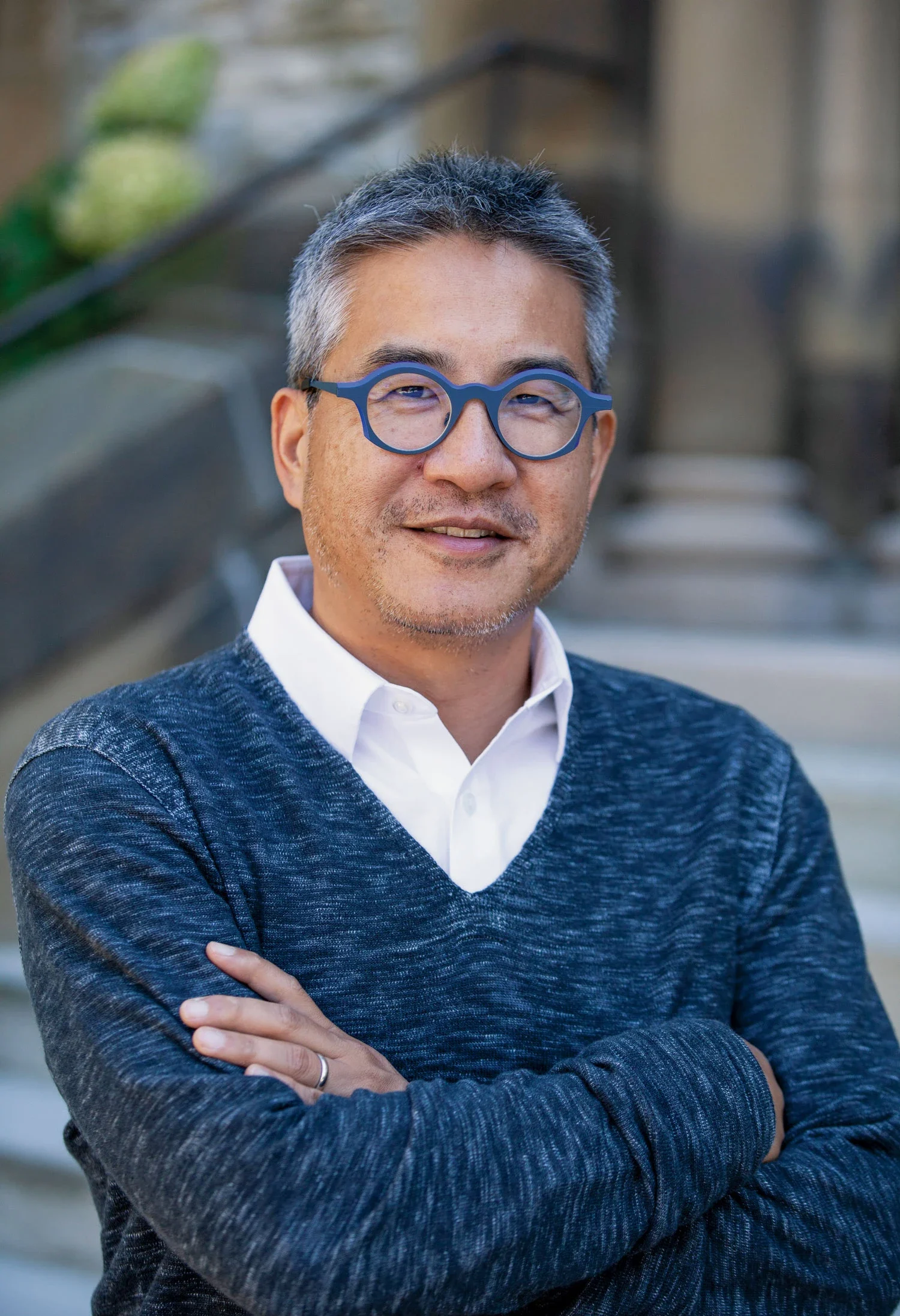 Outdoor photo of Joe Wong in blue framed glasses and V-neck sweater, with his arms crossed