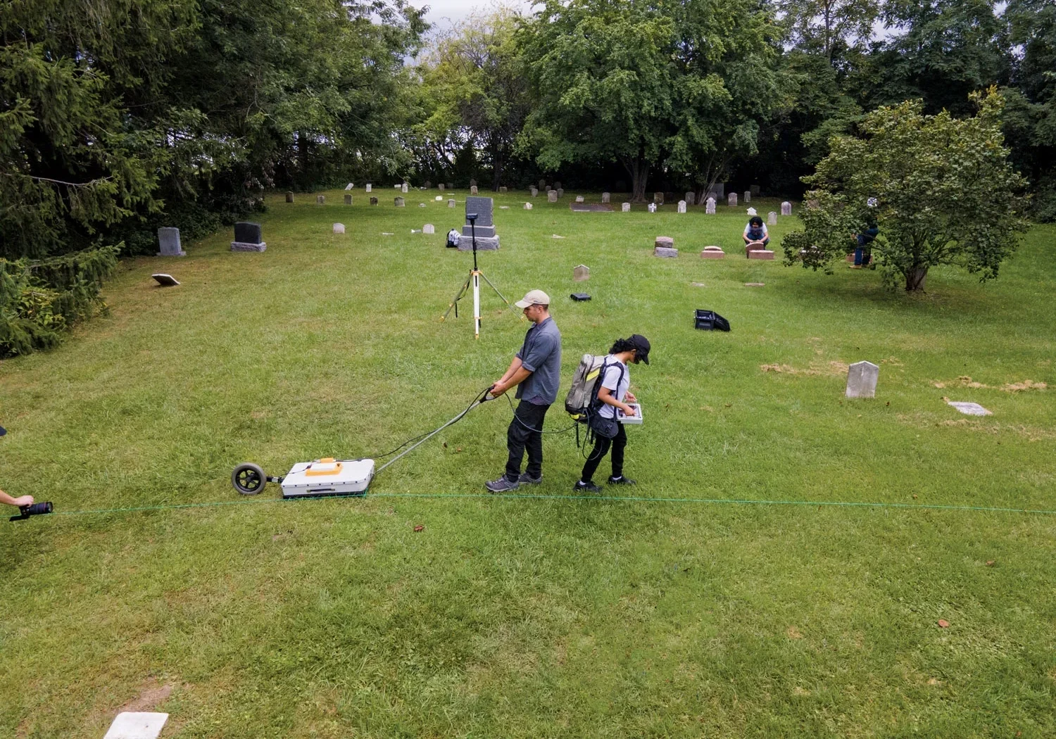 A researcher is pulling a ground-penetrating radar across a cemetery along a marked line. Another researcher is carrying a backpack and operating a monitor.