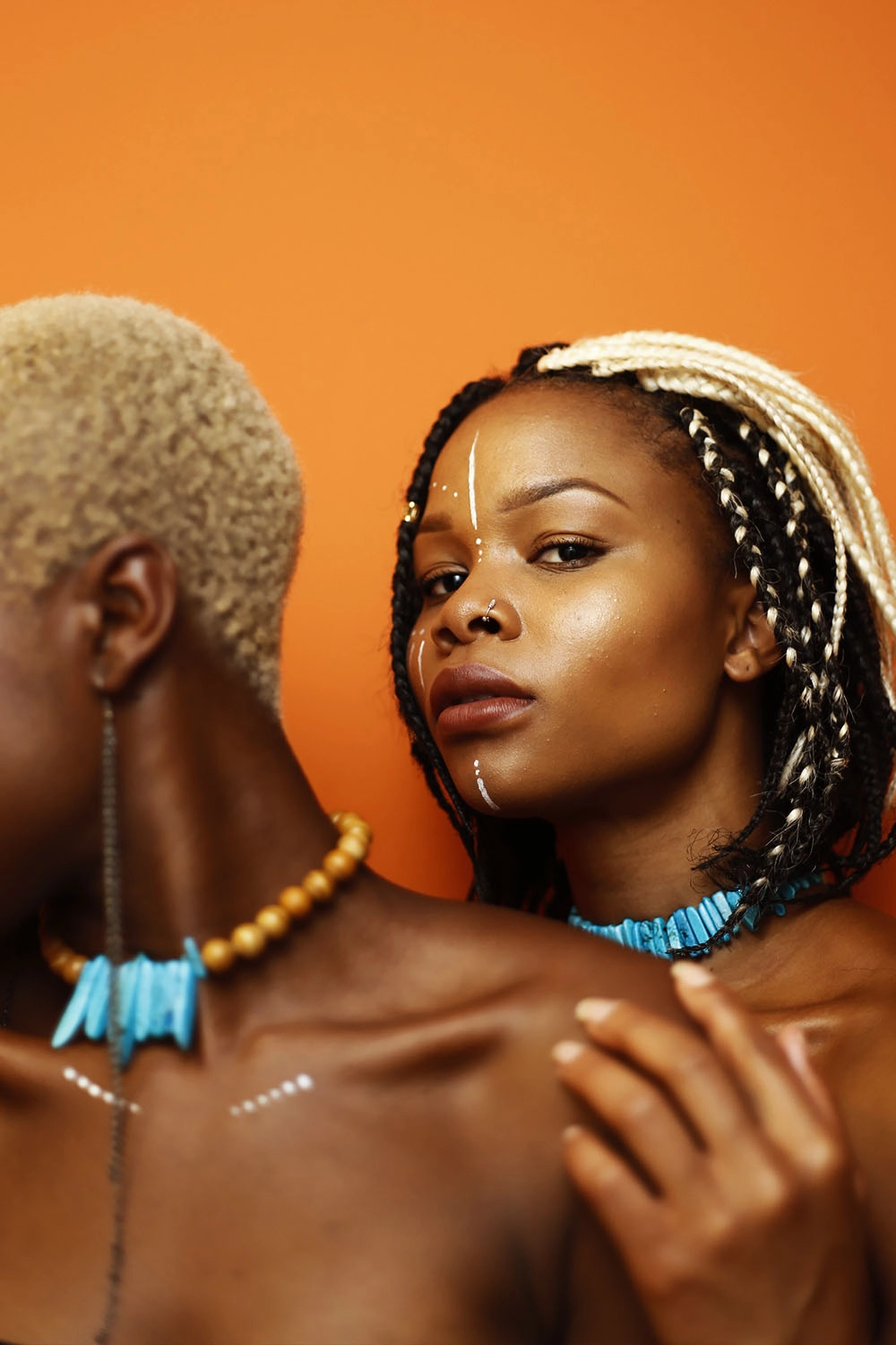 Two Black women against an orange background. One is looking off camera, the other is looking straight ahead and has white face paint and two-toned braids.